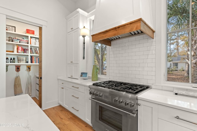 kitchen featuring custom exhaust hood, light stone counters, light hardwood / wood-style flooring, stainless steel stove, and white cabinets