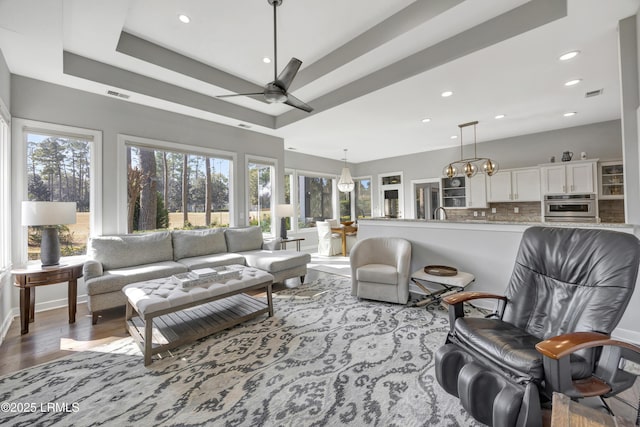 living room with ceiling fan with notable chandelier, light hardwood / wood-style floors, a raised ceiling, and a healthy amount of sunlight