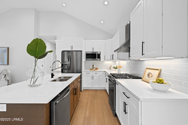 kitchen with wall chimney exhaust hood, a sink, light wood finished floors, appliances with stainless steel finishes, and vaulted ceiling