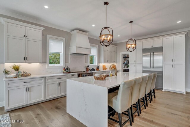 kitchen featuring premium range hood, appliances with stainless steel finishes, an island with sink, white cabinets, and decorative backsplash