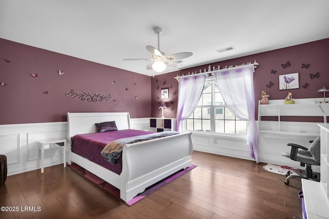 bedroom featuring visible vents, wood-type flooring, a decorative wall, a ceiling fan, and wainscoting
