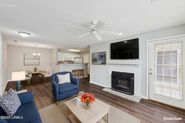 living room with dark wood-style floors, ceiling fan with notable chandelier, a tile fireplace, and baseboards