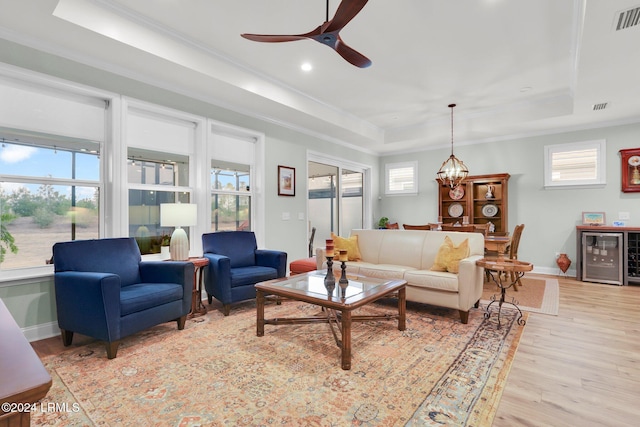 living room with ceiling fan with notable chandelier, beverage cooler, ornamental molding, light hardwood / wood-style floors, and a raised ceiling