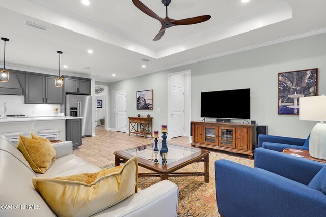 living room featuring sink, crown molding, light wood-type flooring, a tray ceiling, and ceiling fan