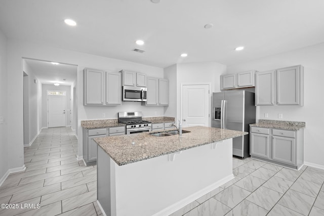 kitchen featuring gray cabinets, sink, light stone counters, stainless steel appliances, and a center island with sink