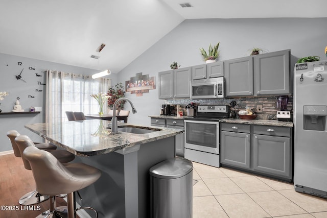 kitchen featuring gray cabinets, appliances with stainless steel finishes, a kitchen island with sink, a sink, and a kitchen breakfast bar