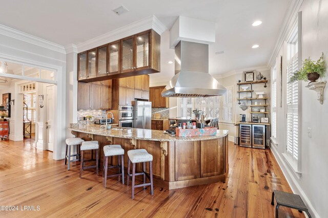 kitchen with island range hood, ornamental molding, kitchen peninsula, stainless steel appliances, and light stone countertops