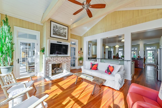 living room with a ceiling fan, dark wood-style floors, beamed ceiling, wood walls, and a brick fireplace