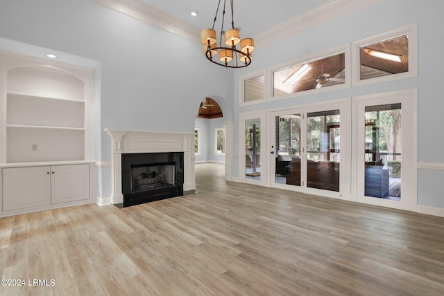 unfurnished living room with a towering ceiling, a notable chandelier, ornamental molding, built in shelves, and light wood-type flooring