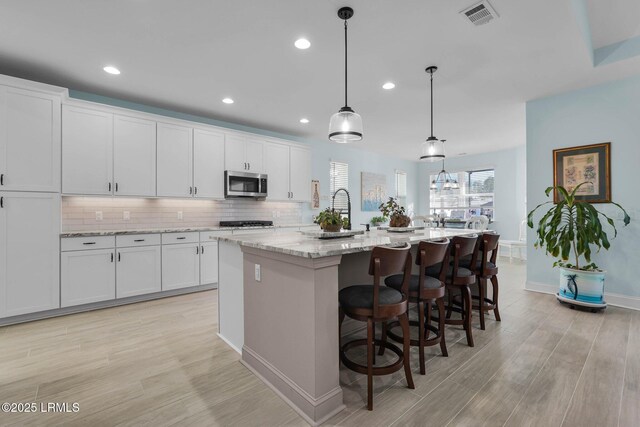 kitchen with light stone counters, an island with sink, hanging light fixtures, and white cabinets