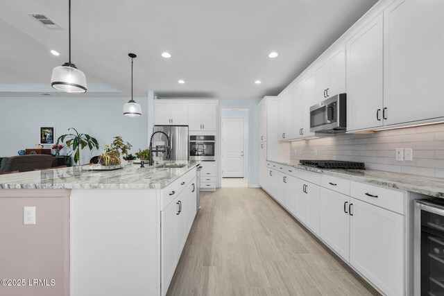 kitchen with appliances with stainless steel finishes, white cabinetry, beverage cooler, hanging light fixtures, and a center island with sink