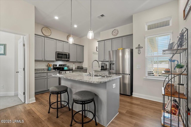 kitchen featuring light stone counters, stainless steel appliances, a sink, visible vents, and a center island with sink