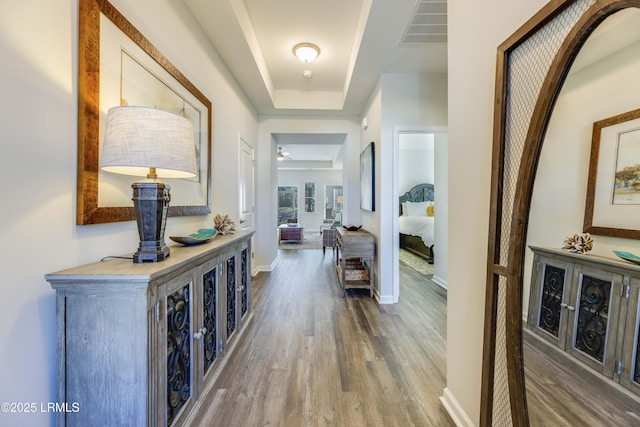 hall featuring baseboards, a tray ceiling, visible vents, and wood finished floors