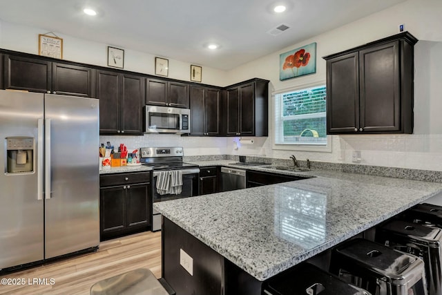 kitchen with dark brown cabinetry, light wood-type flooring, kitchen peninsula, stainless steel appliances, and light stone countertops