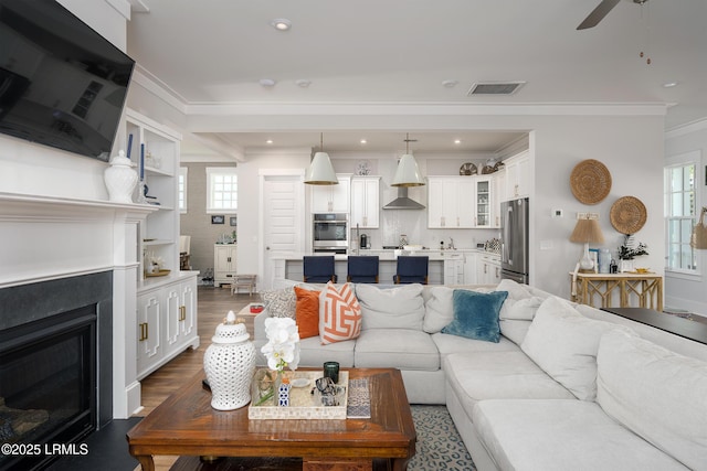 living area with ornamental molding, a glass covered fireplace, ceiling fan, visible vents, and dark wood-type flooring