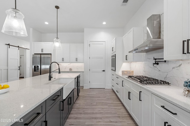 kitchen featuring a barn door, stainless steel appliances, wall chimney exhaust hood, decorative light fixtures, and white cabinetry