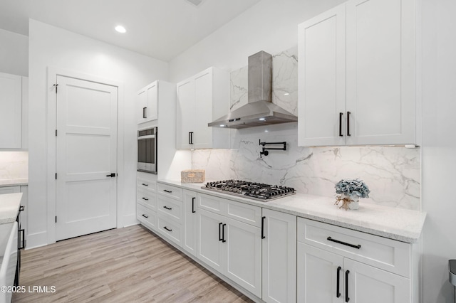 kitchen with wall chimney range hood, decorative backsplash, light wood-style flooring, white cabinets, and appliances with stainless steel finishes