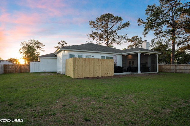 back of house at dusk featuring a lawn, a chimney, a fenced backyard, and a sunroom