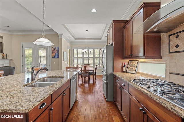 kitchen with a tray ceiling, appliances with stainless steel finishes, a sink, wall chimney range hood, and an island with sink