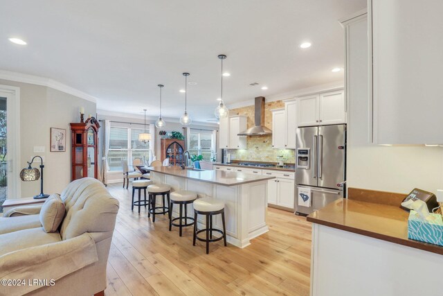 kitchen with white cabinets, high end fridge, wall chimney range hood, and a kitchen island with sink