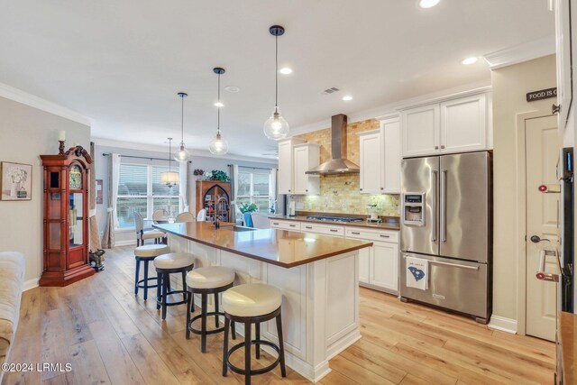 kitchen with appliances with stainless steel finishes, an island with sink, white cabinets, decorative light fixtures, and wall chimney exhaust hood