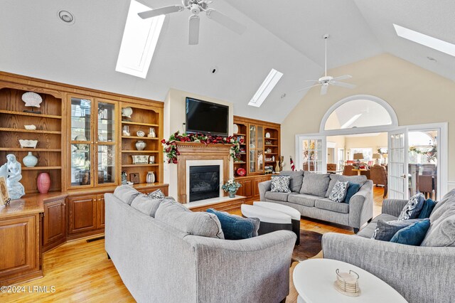 living room with high vaulted ceiling, a skylight, ceiling fan, light wood-type flooring, and french doors