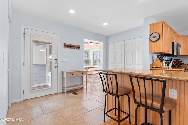 kitchen featuring a breakfast bar, stainless steel microwave, light brown cabinetry, light stone counters, and recessed lighting