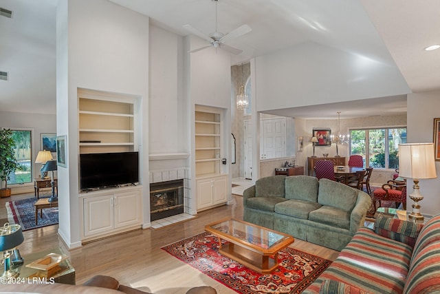 living room with a tile fireplace, ceiling fan with notable chandelier, built in features, high vaulted ceiling, and light hardwood / wood-style floors