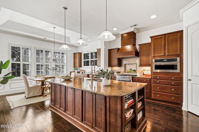 kitchen featuring premium range hood, appliances with stainless steel finishes, hanging light fixtures, a kitchen island with sink, and light stone countertops