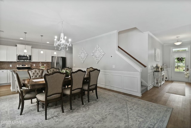dining room with a notable chandelier, crown molding, stairway, light wood-type flooring, and wainscoting