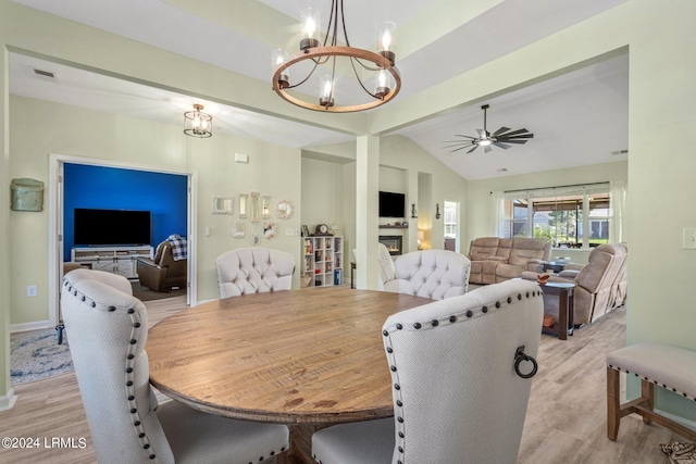 dining space with a warm lit fireplace, lofted ceiling with beams, light wood finished floors, and ceiling fan with notable chandelier