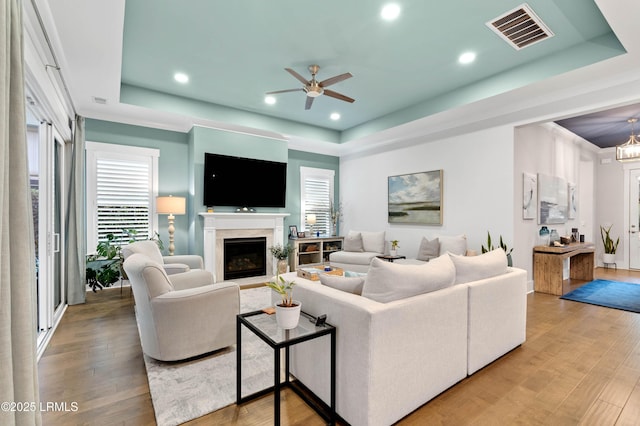 living room with hardwood / wood-style flooring, a high end fireplace, a raised ceiling, and ceiling fan with notable chandelier