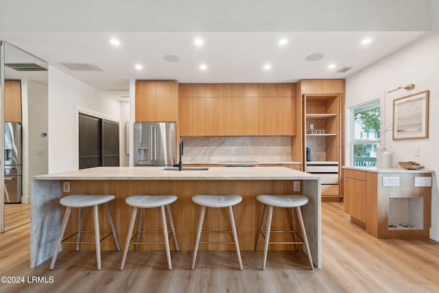 kitchen with stainless steel fridge with ice dispenser, backsplash, light hardwood / wood-style flooring, and an island with sink