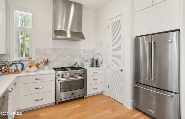 kitchen with wall chimney exhaust hood, white cabinetry, light hardwood / wood-style flooring, premium appliances, and backsplash