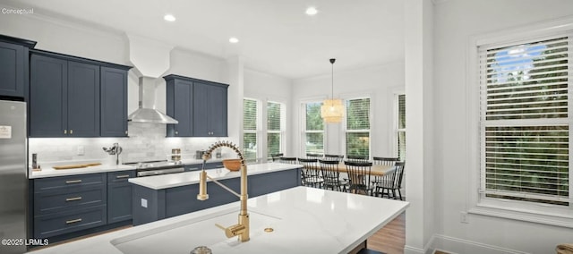 kitchen featuring appliances with stainless steel finishes, decorative backsplash, wall chimney exhaust hood, an island with sink, and pendant lighting