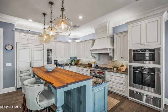 kitchen featuring a center island with sink, appliances with stainless steel finishes, hanging light fixtures, premium range hood, and a warming drawer