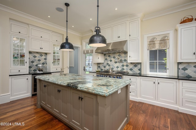 kitchen featuring hanging light fixtures, dark stone countertops, a kitchen island with sink, decorative backsplash, and wall chimney range hood