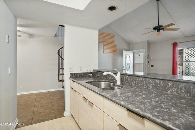 kitchen featuring lofted ceiling with skylight, light brown cabinetry, sink, dark stone countertops, and ceiling fan