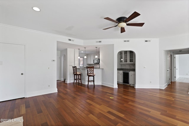 unfurnished living room featuring wine cooler, a ceiling fan, arched walkways, ornamental molding, and dark wood-style floors
