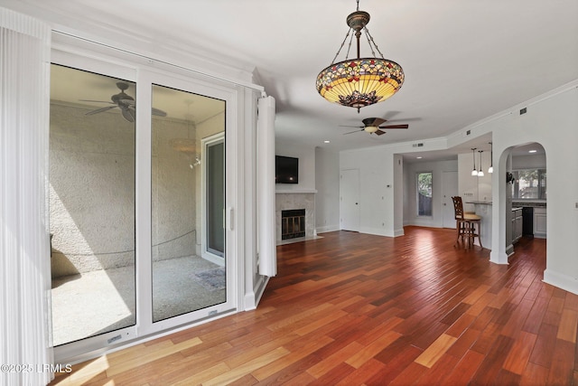 unfurnished living room featuring ceiling fan, a tiled fireplace, arched walkways, wood finished floors, and crown molding