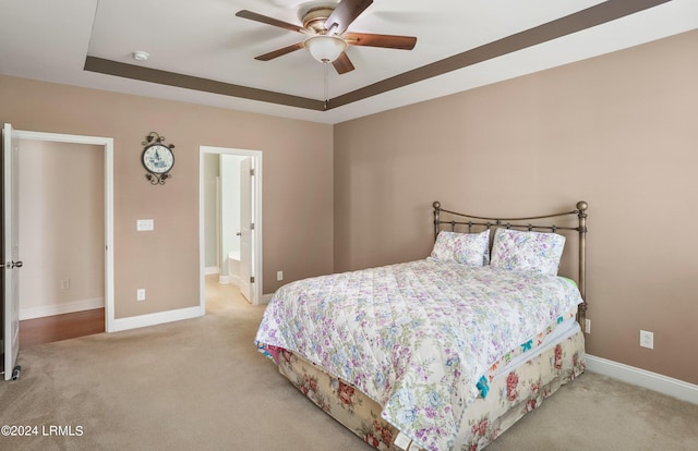 bedroom featuring ceiling fan, ensuite bathroom, a tray ceiling, and light colored carpet