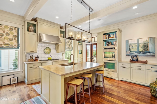 kitchen with appliances with stainless steel finishes, beamed ceiling, light stone countertops, a sink, and exhaust hood