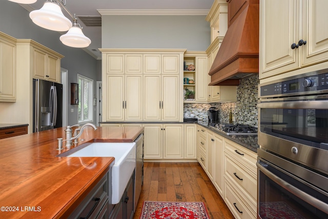 kitchen with butcher block countertops, decorative light fixtures, custom range hood, stainless steel appliances, and cream cabinets