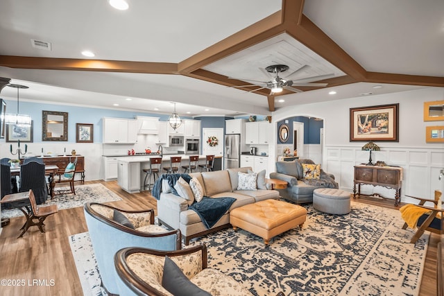 living room featuring a ceiling fan, beam ceiling, light wood-type flooring, visible vents, and arched walkways