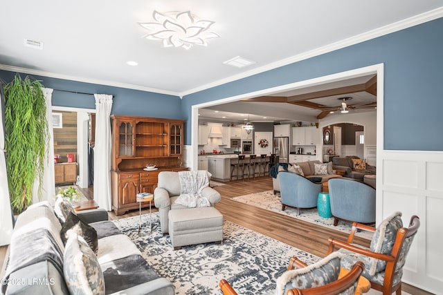 living area with a wainscoted wall, visible vents, crown molding, light wood finished floors, and a ceiling fan