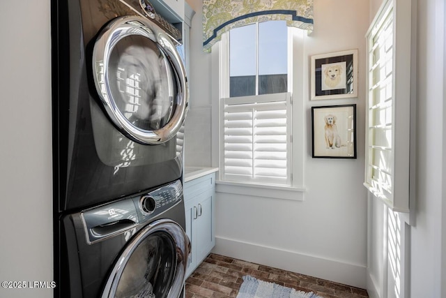 laundry room featuring stacked washing maching and dryer, baseboards, brick floor, and cabinet space