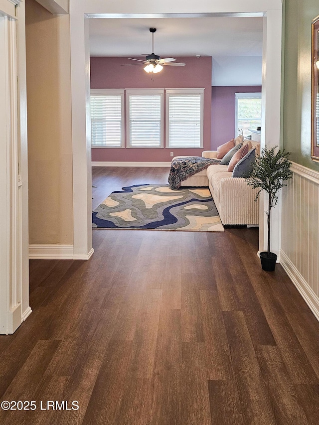 living area with a wainscoted wall, ceiling fan, dark wood-style flooring, and baseboards