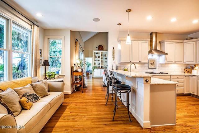 kitchen featuring white cabinets, a kitchen bar, hanging light fixtures, light stone countertops, and wall chimney exhaust hood