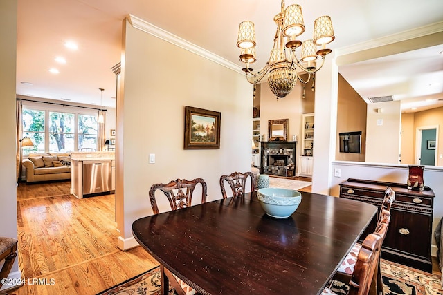 dining room with crown molding, light hardwood / wood-style flooring, built in features, a notable chandelier, and a stone fireplace