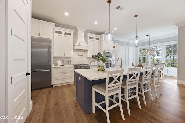 kitchen featuring an island with sink, custom range hood, pendant lighting, stainless steel appliances, and white cabinets